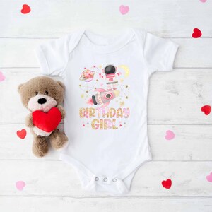 May include: White baby onesie with a colorful space-themed graphic featuring an astronaut, rocket, planets, and the text "BIRTHDAY GIRL". A brown teddy bear holding a red heart is next to the onesie. Scattered red and pink heart-shaped confetti.