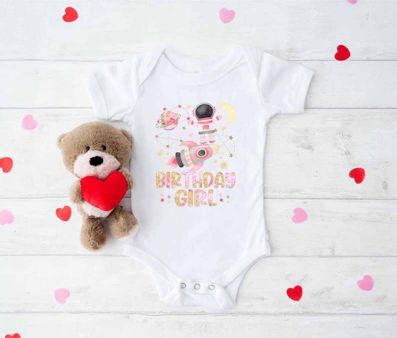 May include: White baby onesie with a colorful space-themed graphic featuring an astronaut, rocket, planets, and the text "BIRTHDAY GIRL". A brown teddy bear holding a red heart is next to the onesie. Scattered red and pink heart-shaped confetti.