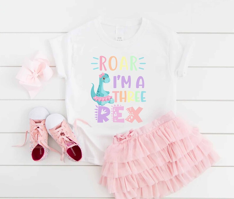 May include: A white t-shirt with a rainbow dinosaur graphic and the text "Roar I'm a Three Rex". The shirt is paired with a pink tulle skirt and pink sparkly shoes.