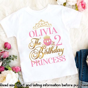 Pu&ograve; includere: Maglietta bianca con un design rosa e oro. Il design presenta una corona, una carrozza e il testo "Olivia The 2nd Birthday Princess".