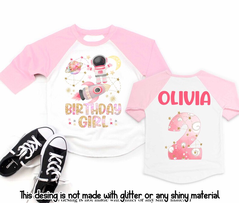 May include: White and pink raglan-style birthday shirt with an astronaut, rocket, and planet design. The shirt says "BIRTHDAY GIRL" in gold glitter lettering. A second shirt shows the name "OLIVIA" and the number "2".