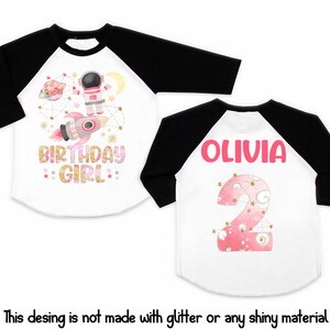 May include: White and black raglan-style birthday shirt. The front features an astronaut, rocket, and the words "BIRTHDAY GIRL." The back displays the name "OLIVIA" and a pink number 2. The design is not made with glitter.