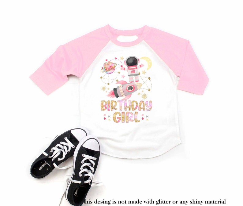 May include: White and pink raglan-style t-shirt with a space-themed graphic. The design includes an astronaut, rocket, planets, and the text "BIRTHDAY GIRL". Black and white sneakers are at the bottom of the image.