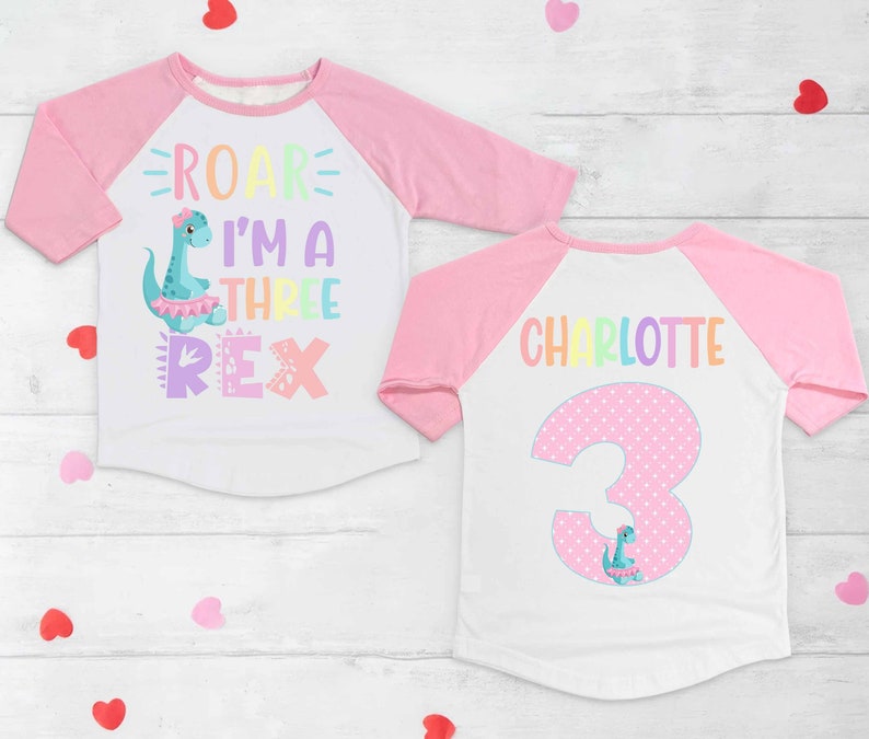 May include: A pink and white long-sleeved shirt with a rainbow dinosaur graphic and the text "Roar I'm a Three Rex". The back of the shirt has a pink polka dot number 3 with a dinosaur graphic and the name "Charlotte".