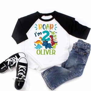May include: A white and black baseball tee shirt with a dinosaur graphic and the text "Roar I'm 2 Oliver".
