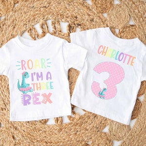 May include: Two white t-shirts with a dinosaur theme. One shirt says "Roar I'm a Three Rex" with a pink, blue, and green dinosaur. The other shirt says "Charlotte" in rainbow letters and has a pink number 3 with a small dinosaur.