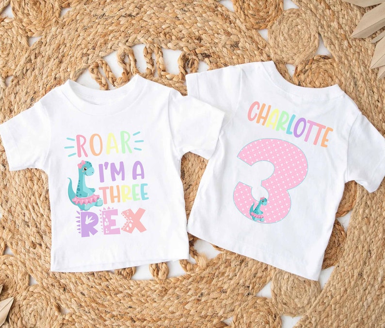 May include: Two white t-shirts with a dinosaur theme. One shirt says "Roar I'm a Three Rex" with a pink, blue, and green dinosaur. The other shirt says "Charlotte" in rainbow letters and has a pink number 3 with a small dinosaur.