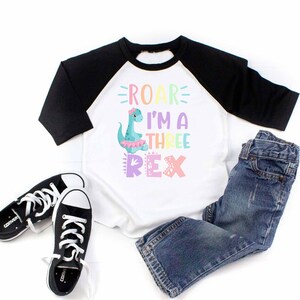 May include: A white and black baseball tee shirt with a colorful dinosaur graphic and the text "ROAR I'M A THREE REX". The shirt is paired with blue jeans and black sneakers.