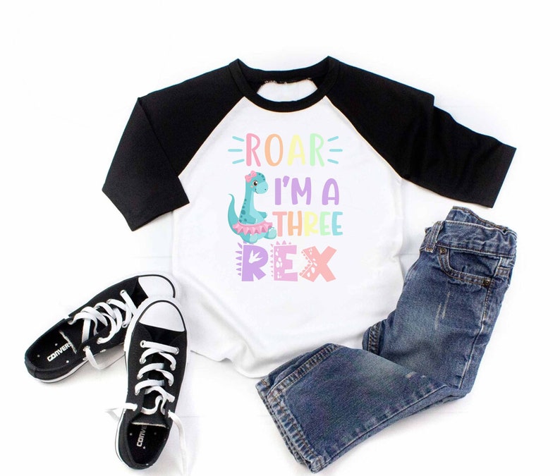 May include: A white and black baseball tee shirt with a colorful dinosaur graphic and the text "ROAR I'M A THREE REX". The shirt is paired with blue jeans and black sneakers.