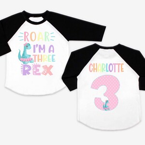May include: Two white and black baseball shirts with a dinosaur theme. One shirt says "Roar I'm a Three Rex" with a pink dinosaur. The other shirt says "Charlotte" with a pink number 3 and a small dinosaur.