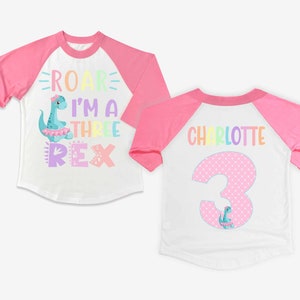 May include: A white and pink baseball style t-shirt with a rainbow dinosaur graphic and the text "Roar I'm a Three Rex". The back of the shirt has a pink polka dot number 3 with a dinosaur graphic and the name "Charlotte" in rainbow letters.