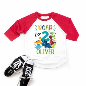 May include: A white and red baseball tee shirt with a dinosaur theme. The shirt says "Roar I'm 2 Oliver".
