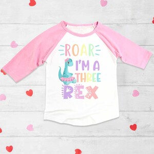 May include: A white and pink long-sleeved shirt with a rainbow dinosaur graphic and the text "Roar I'm a Three Rex".