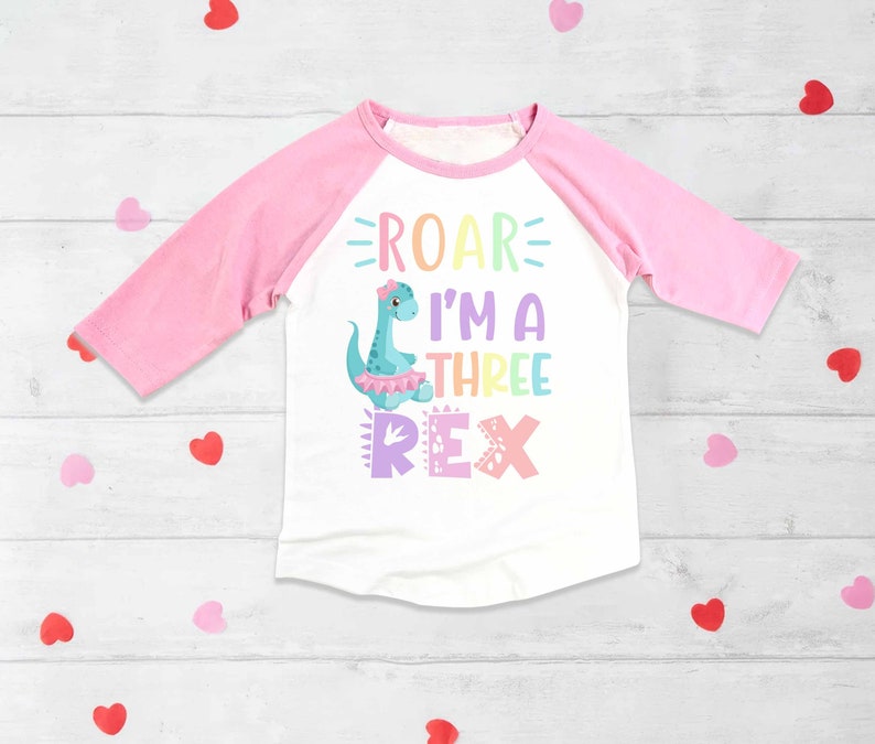 May include: A white and pink long-sleeved shirt with a rainbow dinosaur graphic and the text "Roar I'm a Three Rex".