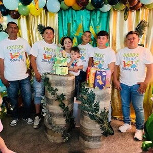May include: A group of men and children wearing white t-shirts with dinosaur graphics and the text "Cousin of the Birthday Boy", "Uncle of the Birthday Boy", "Brother of the Birthday Boy", "Uncle of the Birthday Boy", and "Daddy of the Birthday Boy". The men are standing in front of a green and gold backdrop with balloons and a dinosaur-themed cake.