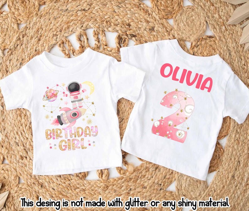 May include: Two white t-shirts with colorful designs. One features an astronaut, rocket, planets, and the text "BIRTHDAY GIRL". The other has the name "OLIVIA" and a pink number "2" with stars. The text at the bottom says "This desing is not made with glitter or any shiny material."