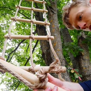 May include: A close-up of a wooden ladder made of rope and wooden rungs. The ladder is being held by a person's hand. The ladder is hanging from a tree branch.