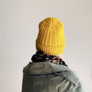 Może przedstawiać: Żółty wełniany beret z fakturowanym wzorem. Beret nosi osoba w zielonej kurtce i czarno-białym wzorzystym szaliku.