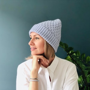 Könnte beinhalten: Eine Frau trägt eine helllilafarbene Strickmütze mit einem strukturierten Muster. Sie trägt ein weißes Hemd mit Knöpfen und hat die Hand am Kinn.