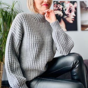 May include: A gray knitted sweater with a mock neck and long sleeves. The sweater has a ribbed texture and is worn with black leather pants.