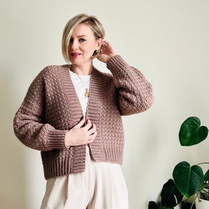 Könnte beinhalten: Eine altrosa gehäkelte Strickjacke mit strukturiertem Maschenmuster, langen Ärmeln mit gerippten Bündchen und einer breiten, gerippten Knopfleiste. Das Kleidungsstück wird über einem schlichten weißen T-Shirt und einer hellen Hose getragen.
