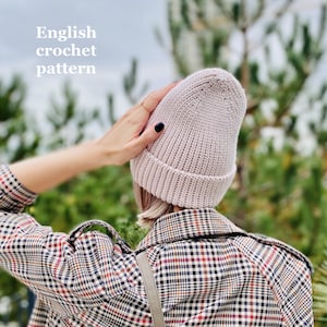 Könnte beinhalten: Nahaufnahme einer rosafarbenen Häkelmütze. Die Mütze wird von einer Person getragen, die ihre Hand auf die Mütze legt. Die Person trägt ein kariertes Hemd.