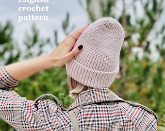 May include: A close-up of a pink crocheted beanie hat. The hat is being worn by a person with their hand on top of the hat. The person is wearing a plaid shirt.