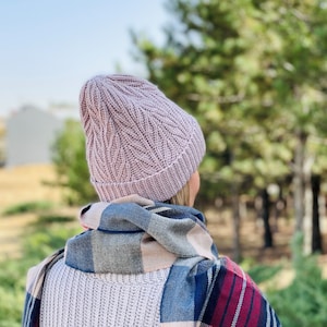 Op de afbeelding: Een vrouw draagt ​​een roze gebreide muts met een kabelbreipatroon en een geruite sjaal. De muts is over haar oren getrokken en ze kijkt weg van de camera.