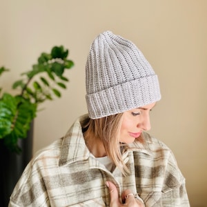 Könnte beinhalten: Eine hellgraue Häkelmütze mit Rippenstruktur. Die Mütze wird von einer Person mit langen blonden Haaren und einem karierten Hemd getragen.