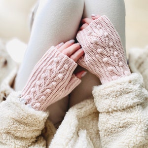 May include: A pair of pink fingerless gloves with a textured stitch pattern and small white bobbles. The gloves are made of a soft yarn and have a delicate, feminine look.