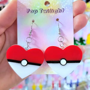 May include: Heart-shaped earrings with a red and white design, featuring a black stripe and a white circle. The earrings are on a card that says "Pop Twilight".