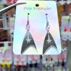 May include: Silver-coloured earrings in a Star Trek insignia shape, with a star in the centre. The earrings hang from a silver hook. The earrings are displayed on a Pop Twilight card.