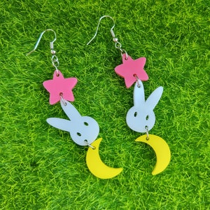 Puede incluir: Pendientes colgantes con un dise&ntilde;o de estrella rosa, conejo blanco y media luna amarilla. Los pendientes son de acr&iacute;lico y tienen ganchos plateados. El dise&ntilde;o es caprichoso y juguet&oacute;n, perfecto para a&ntilde;adir un toque de diversi&oacute;n a cualquier atuendo.