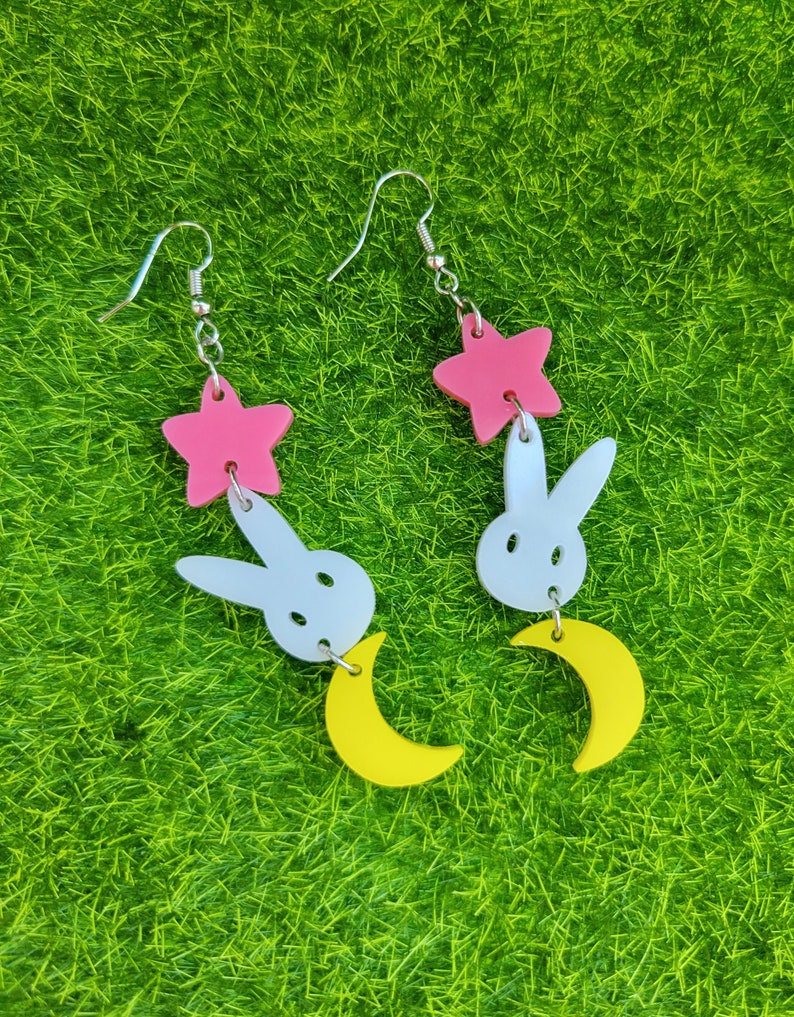 Puede incluir: Pendientes colgantes con un dise&ntilde;o de estrella rosa, conejo blanco y media luna amarilla. Los pendientes son de acr&iacute;lico y tienen ganchos plateados. El dise&ntilde;o es caprichoso y juguet&oacute;n, perfecto para a&ntilde;adir un toque de diversi&oacute;n a cualquier atuendo.