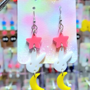 Puede incluir: Pendientes con un dise&ntilde;o de estrella rosa, conejo blanco y luna creciente amarilla. Los pendientes cuelgan de una tarjeta de exhibici&oacute;n que dice "Pop Twilight". Los pendientes son de acr&iacute;lico.