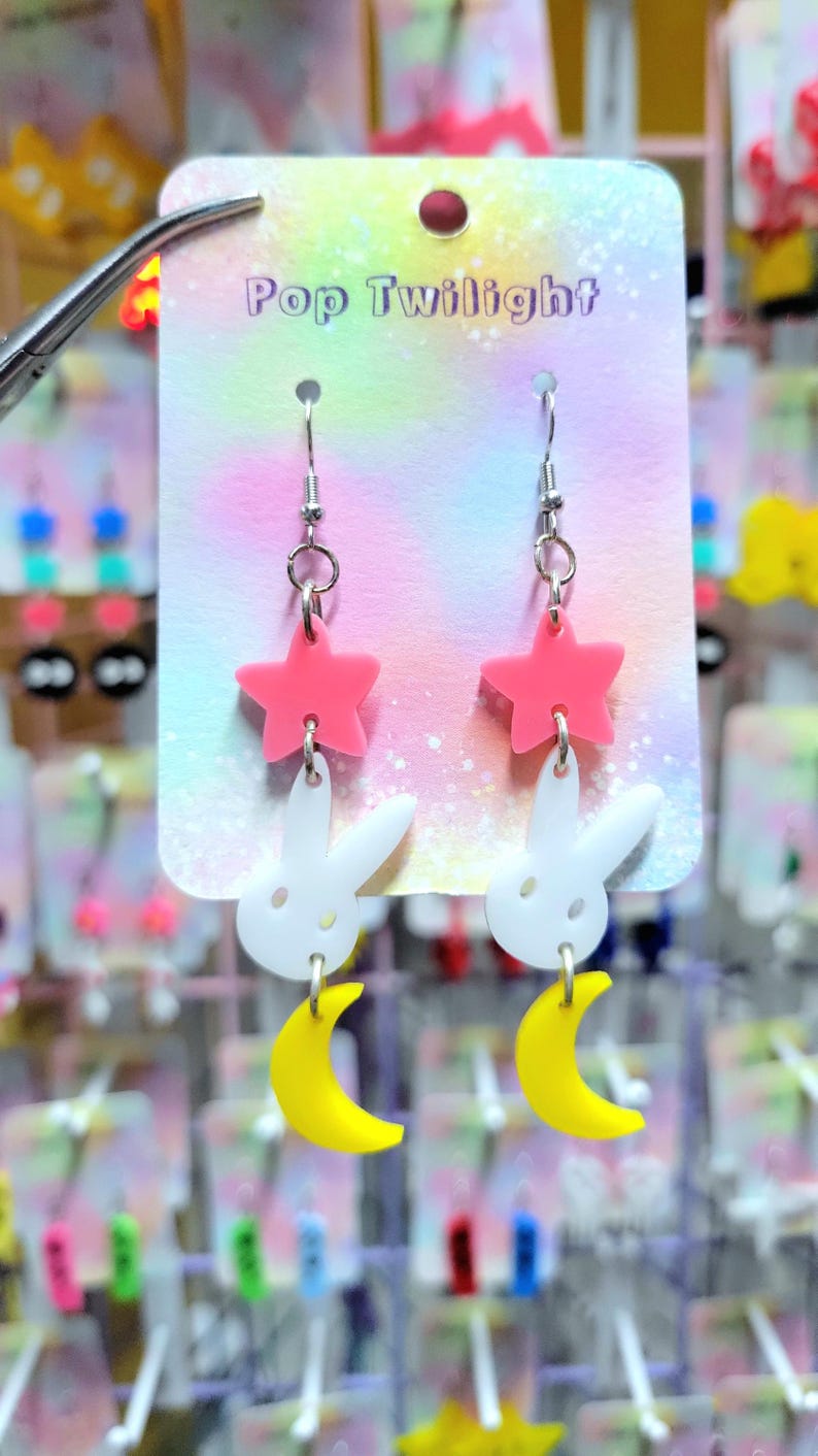 Puede incluir: Pendientes con un dise&ntilde;o de estrella rosa, conejo blanco y luna creciente amarilla. Los pendientes cuelgan de una tarjeta de exhibici&oacute;n que dice "Pop Twilight". Los pendientes son de acr&iacute;lico.