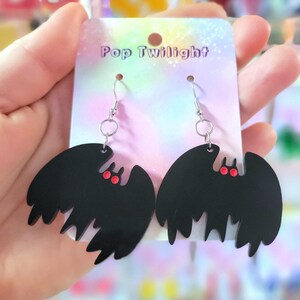 Puede incluir: Pendientes negros con forma de murciélago y ojos rojos, colgados de ganchos plateados. Los pendientes se muestran en una tarjeta que dice "Pop Twilight". Los pendientes tienen un borde inferior dentado.