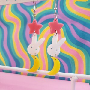 Puede incluir: Un par de pendientes colgantes con un dise&ntilde;o caprichoso. Cada pendiente presenta una estrella rosa, un conejito blanco y una luna creciente amarilla. Los pendientes est&aacute;n suspendidos de ganchos plateados, creando un accesorio l&uacute;dico y &uacute;nico.