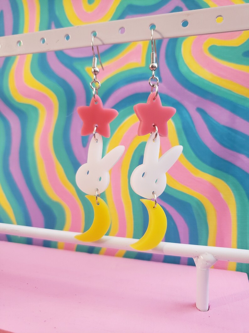 Puede incluir: Un par de pendientes colgantes con un dise&ntilde;o caprichoso. Cada pendiente presenta una estrella rosa, un conejito blanco y una luna creciente amarilla. Los pendientes est&aacute;n suspendidos de ganchos plateados, creando un accesorio l&uacute;dico y &uacute;nico.