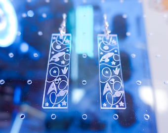 Pode incluir: Brincos retangulares transparentes com desenhos gravados. Cada brinco apresenta uma nave espacial, estrelas e linhas onduladas. Os brincos pendem de ganchos prateados. O fundo é azul desfocado.
