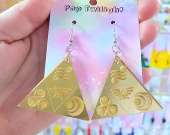 Pode incluir: Brincos triangulares dourados com desenhos gravados, segurados em frente a uma placa Pop Twilight. Os brincos têm fecho de gancho e apresentam um brasão estilizado e outros símbolos. O fundo é uma exibição de vários acessórios coloridos.