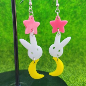 Puede incluir: Pendientes colgantes con una estrella rosa, un conejito blanco y una luna creciente amarilla. Los pendientes cuelgan de ganchos plateados. El fondo es de color verde brillante.