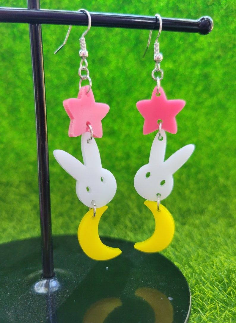 Puede incluir: Pendientes colgantes con una estrella rosa, un conejito blanco y una luna creciente amarilla. Los pendientes cuelgan de ganchos plateados. El fondo es de color verde brillante.