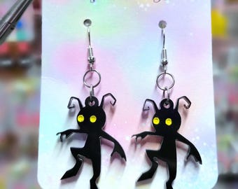 Pode incluir: Brincos de silhueta preta com olhos amarelos, apresentando uma figura estilizada com braços e pernas estendidos. Os brincos penduram de ganchos prateados. O cartão de apoio diz "Pop Twilight" em texto roxo.