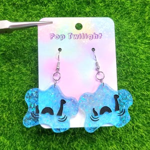 Puede incluir: Un par de pendientes azules en forma de estrella con detalles de purpurina. Los pendientes tienen un diseño de cara de dibujos animados y están suspendidos de ganchos plateados. El fondo es verde.