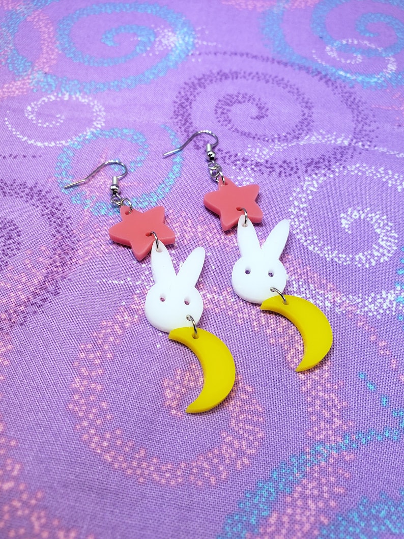 Puede incluir: Un par de pendientes caprichosos con una estrella rosa, un conejito blanco y una luna creciente amarilla. Los pendientes son de acr&iacute;lico y tienen ganchos plateados. El dise&ntilde;o es juguet&oacute;n y colorido.