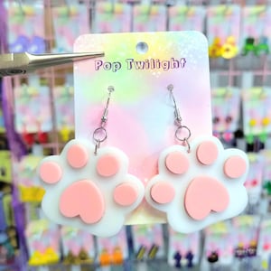 Può includere: Orecchini a forma di zampa bianchi e rosa con ganci color argento. Gli orecchini sono esposti su una carta con la scritta "Pop Twilight". Lo sfondo presenta una varietà di altri orecchini.