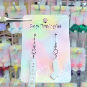 Puede incluir: Pendientes con un mando a distancia blanco y una forma abstracta blanca. Los pendientes están en una tarjeta que dice "Pop Twilight". Se ven otros pendientes con varias formas en el fondo.