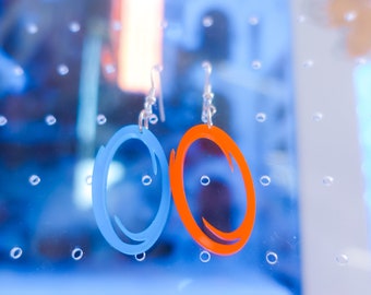 Pode incluir: Um par de brincos com um design de portal azul e laranja. Os brincos são pendurados em ganchos prateados. O design é uma forma oval com uma abertura curva. São feitos de um material leve.