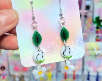Pode incluir: Brincos pendurados com tema floral. Cada brinco apresenta uma folha verde, um botão de flor branco e uma pequena flor branca com centro amarelo. A inscrição "Pop Twilight" é visível no cartão.